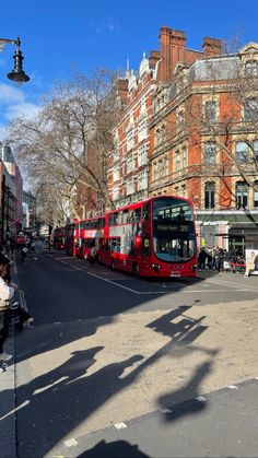 a red double decker bus driving down a street next to tall buildings and people sitting on benches