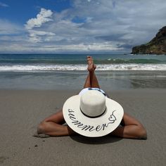 a woman laying on top of a sandy beach next to the ocean wearing a white hat