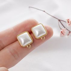 a pair of white and gold square shaped earrings on someone's finger with cherry blossoms in the background