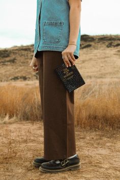 a woman standing on top of a dry grass field holding a black purse and looking at the ground