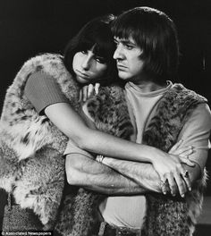 two people standing next to each other with their arms around one another, both wearing fur vests