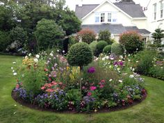 a flower garden in front of a white house with lots of flowers around the perimeter