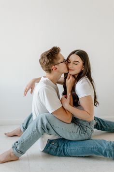 a young man and woman sitting on the floor kissing each other with their arms around one another