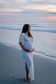 a pregnant woman standing on the beach at sunset wearing a white dress and holding her belly