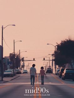 two people are walking down the street in front of some cars and traffic lights at sunset