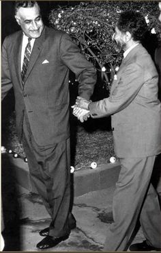 two men shaking hands in front of a tree with white balls all over it and one man wearing a suit