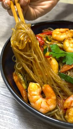 a person holding chopsticks over a bowl of shrimp and noodle stir fry