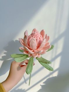 a hand holding a crocheted pink flower with green leaves in the sun light