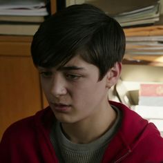 a young man in a red hoodie looking at his cell phone while standing next to a bookshelf