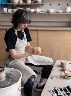 a woman in an apron is sitting on a table with pottery and other items around her