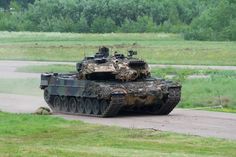 an army tank driving down a road next to a lush green field with trees in the background