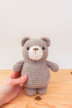 a hand holding a crocheted teddy bear on top of a wooden table next to a white wall