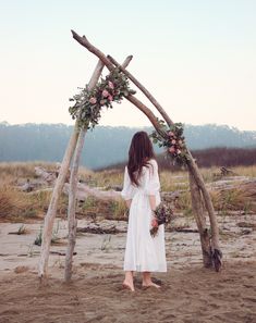 a woman in a white dress standing next to a wooden arch with flowers on it