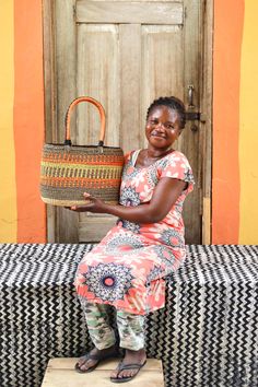 a woman sitting on a bench holding a basket in front of an orange wall and wooden door