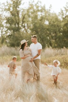 a pregnant couple and their two children are walking through the tall grass in an open field