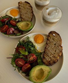 two white plates topped with toast, avocado and tomatoes next to a cup of cappuccino