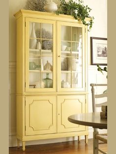 a yellow china cabinet with glass doors and plants on top, in a dining room