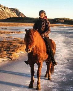 a woman riding on the back of a brown horse across snow covered ground with mountains in the background