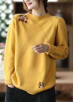 a woman wearing a yellow sweater with flowers on the side and black pants, standing in front of a table