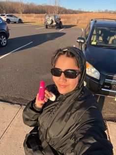 a woman in sunglasses and a black jacket is holding a pink object near a parking lot