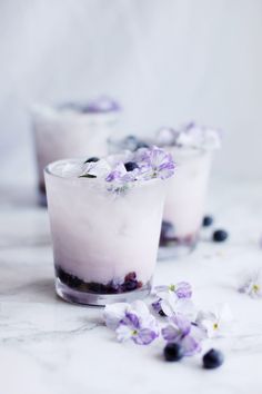 two glasses filled with ice and blueberries on top of a marble table next to flowers