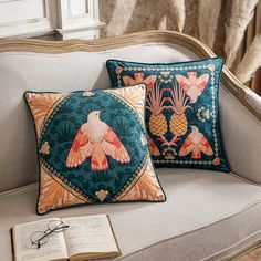 two decorative pillows sitting on top of a couch next to an open book and eyeglasses