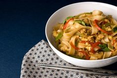 a white bowl filled with noodles and vegetables on top of a blue table cloth next to chopsticks