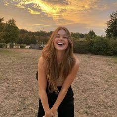 a girl is laughing in the middle of a field with trees and clouds behind her