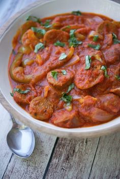 a white bowl filled with meat and tomato sauce on top of a wooden table next to a spoon