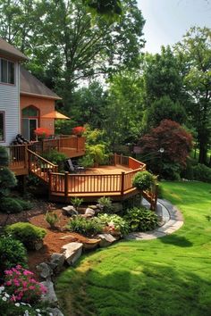 a deck in the middle of a yard with flowers and trees around it, next to a house