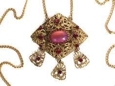 Czech Necklace with Pink Moon Stone Art Glass This exquisite necklace is from the 1920s or 1930s and is Czech. This piece is made of layers of beautiful ornate gold tone filigree in a large diamond shaped pendant. The center stone is a large oval pink cabochon of Czech art glass moon stone! The stone is gorgeous and glows from pink to purple catching the light in magically way. The center stone is surrounded by 8 deep garnet red rhinestones. The final touch is that there are 3 lovely filigree pendants each with  a garnet rhinestone hanging and dangling from the main pendant.  This piece is supported by a long gold chain which closes with a simples ring clasp. Chain Measures 23" long.  Pendant with dangles measures 2 3/8" x 2". This piece is in excellent vintage condition all metal and ston German Jewelry, Czech Jewelry, Filigree Necklaces, Silver Caps, Filigree Pendant, Pink Moon, Red Rhinestone, Moon Stone, Stone Art