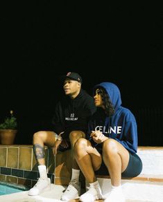 two people sitting next to each other near a swimming pool at night, one wearing a hoodie and the other in shorts