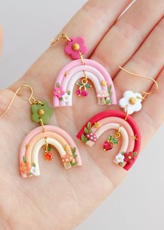 four small rainbow earrings are shown on someone's hand, with flowers in the background