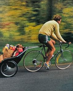 a man riding a bike next to a child in a wagon on the road with trees in the background