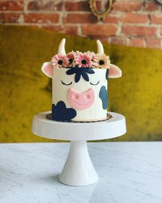 a cake decorated with flowers and a cow face on a white pedestal in front of a brick wall