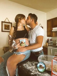 a man and woman kissing while sitting on top of a kitchen counter with food items