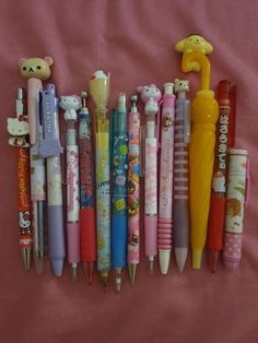 many different types of pens lined up on a pink sheet with teddy bears and other items