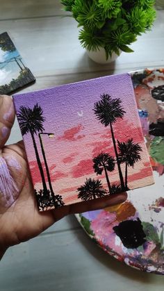 a person holding up a small painting with palm trees on it