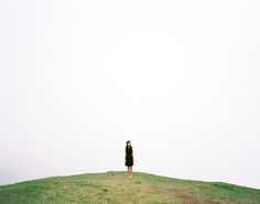 a woman standing on top of a grass covered hill next to a kite flying in the sky