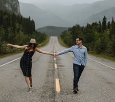 a man and woman holding hands while walking down the middle of an empty road with mountains in the background