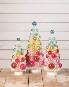 three small christmas trees with colorful ornaments on them sitting on a wooden table in front of a white wall