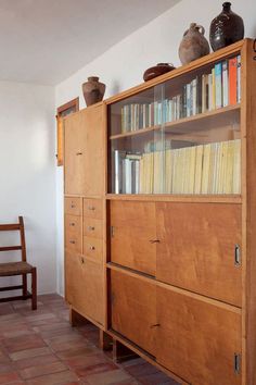 a wooden cabinet with books and vases on it's top shelf next to a chair