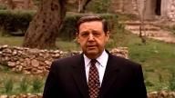 a man wearing a suit and tie standing in front of a stone wall with trees