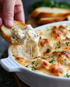 a hand holding a piece of bread and dipping it into a casserole dish