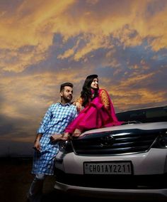 a man and woman standing next to a car under a cloudy sky with the sun setting behind them