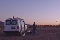 a man standing next to a white van in the middle of a dirt field at sunset