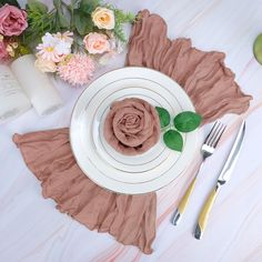 a white plate topped with a pink rose next to two forks and a vase filled with flowers