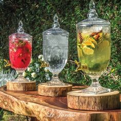 three glasses filled with different types of drinks sitting on top of a wooden table next to bushes