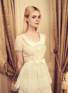 a woman in a white dress standing next to a window with drapes on it