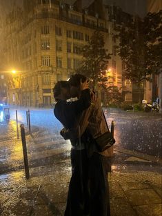 two people kissing in the rain on a city street at night with buildings behind them